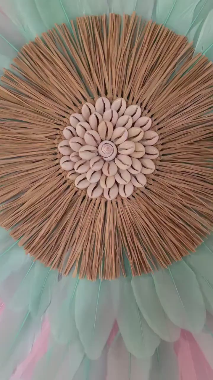 Le RYM :   jujuHat mural en plumes rose tendre et vert d’eau, cœur en cauris et raphia naturel